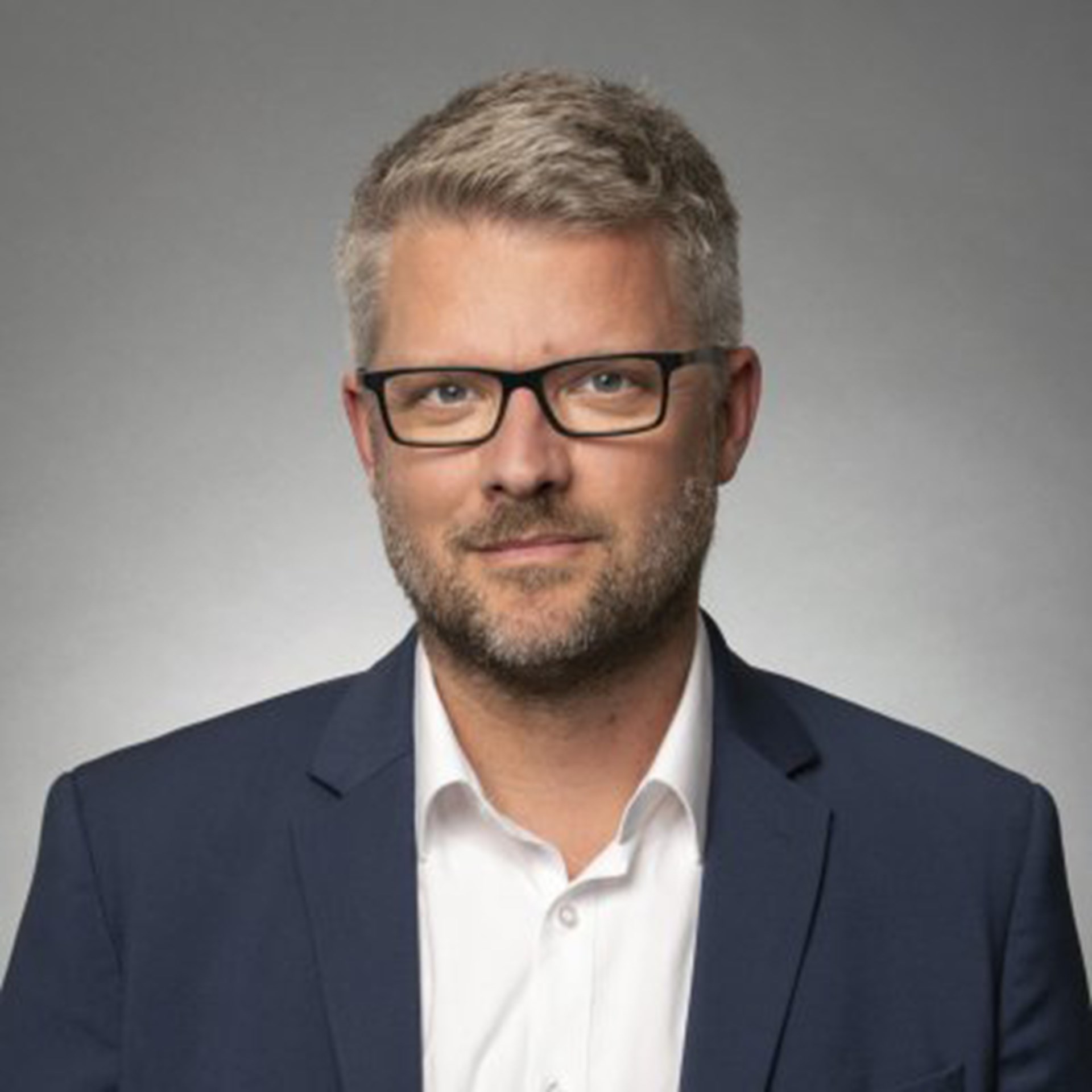 Lächelnder Mann mit blonden Haaren, Brille und Bart, der ein weißes Hemd und eine blaue Anzugjacke trägt