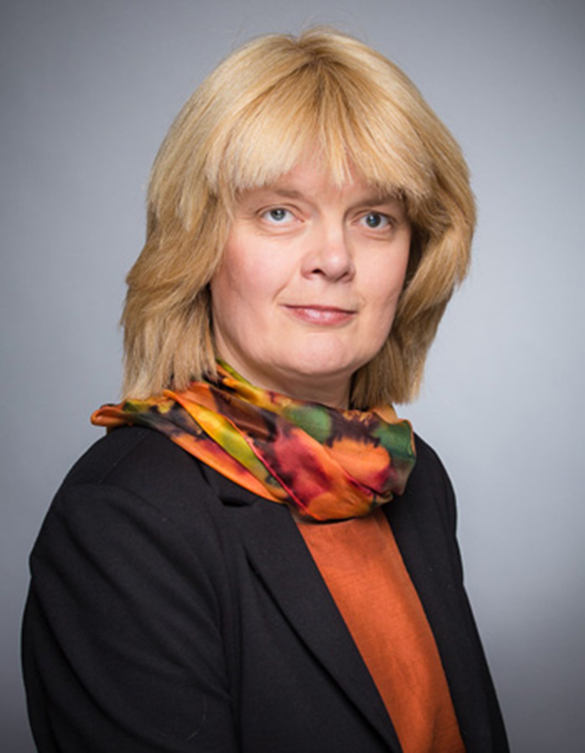 Frau mit schulterlangen, blonden Haaren, die einen bunten Schal, ein orangenes T-Shirt und eine dunkle Anzugjacke trägt